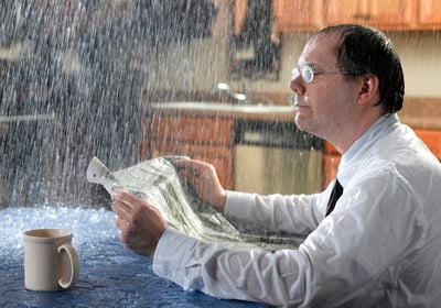 a man sitting at his kitchen table soaking wet reading a paper and drinking coffee while his roof leaks 