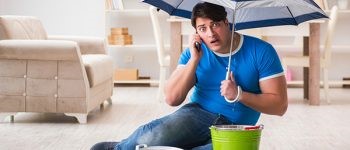 man holding an umbrella inside with a bucket catching roof leaks