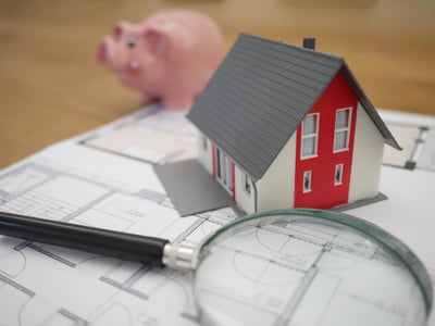 Magnify glass, model home, and piggy bank sitting on blueprints on a table  