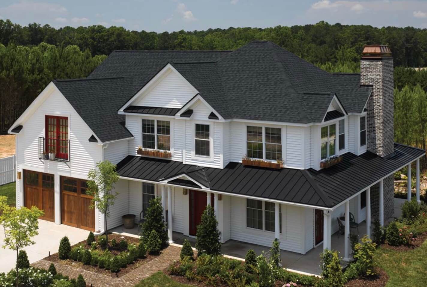 White two-story house with  black shingles and a one story wrap around porch with a metal roof